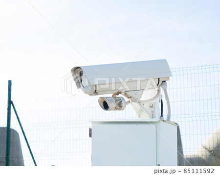 White surveillance camera on a stand outdoors behind a fence White surveillance camera on a stand outdoors behind a fence 85111592