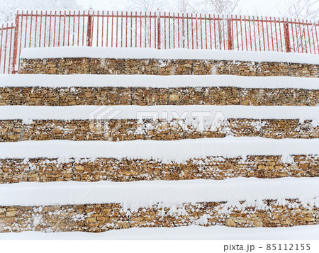 Stepped wall made of stones fixed with a grid with a fence at the top covered with snow Stepped wall made of stones fixed with a grid with a fence at the top covered with snow 85112155