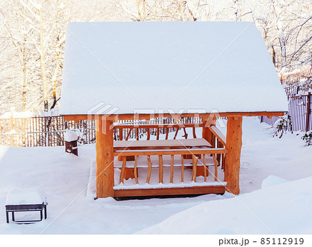 A wooden gazebo covered with snow stands on a snow-covered meadow among trees A wooden gazebo covered with snow stands on a snow-covered meadow among trees 85112919