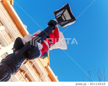 A toy santa claus hangs on a lamppost with a pile of snow on his head against a background of blue sky and building. Bottom view 85113010