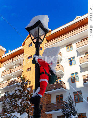 Toy santa claus hanging on a lamppost with a pile of snow on his head against the background of the hotel building 85113011
