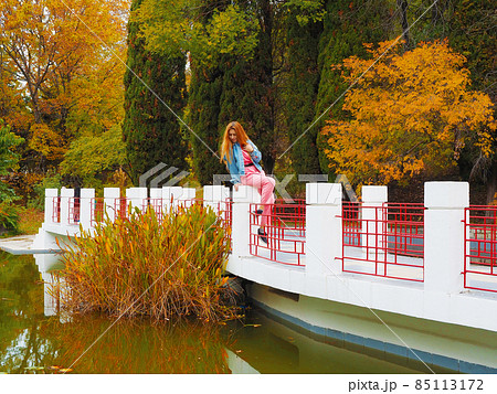 Redhead girl sits on the railing of the bridge over the pond and looks at the water in the park 85113172