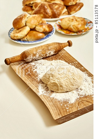 Wooden board with raw dough flour and rolling pin near homemade pies in kitchen Wooden board with raw dough flour and rolling pin near homemade pies in kitchen 85113578