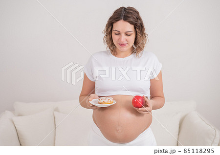 A pregnant woman is holding a red apple and donut. Choice of food. 85118295