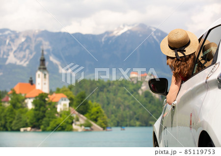 Happy young woman on vacation leaning out of car window on shore of Bled lake, country of Slovenia. Travel, holiday, tourism, explore, rent a car concept. 85119753