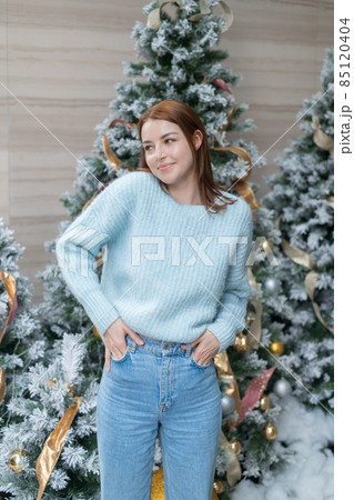 New Year portrait of beautiful smiling caucasian young woman in cozy wool warm light blue sweater standing near decorated Christmas tree. happy person 85120404