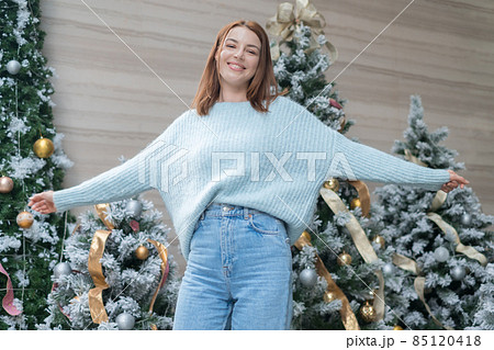 New Year portrait of beautiful smiling caucasian young woman in cozy wool warm light blue sweater standing near decorated Christmas tree. happy person 85120418