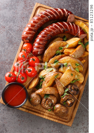 Grilled sausages with potato wedges and mushrooms close-up on a wooden tray. vertical top view 85123296