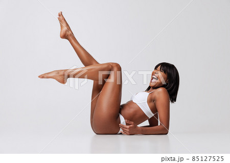 Young cheerful dark skinned girl in white underwear laughing isolated over gray studio background. Natural beauty concept. Young cheerful dark skinned girl in white underwear laughing isolated over gray studio background. Natural beauty concept. 85127525