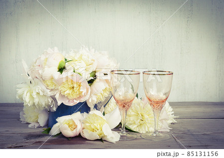Beautiful celebration composition: Two beautiful empty wineglass, bouquet of white peonies on rustic wooden background; copy space 85131156