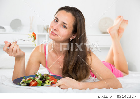 Positive pretty woman holding fork and eating vegetable salad in bed 85142327