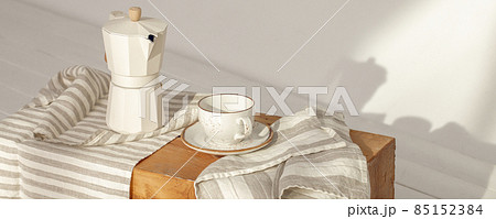 Italian coffee maker with wooden handle and white coffee craft cup on solid wood stamp decorated with tea towels in creamy tones on sunlight creating beautiful shadows on the wall 85152384