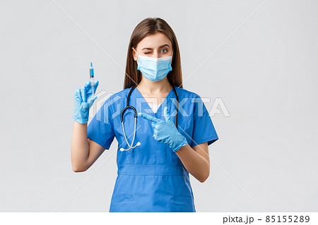 Covid-19, preventing virus, health, healthcare workers and quarantine concept. Cute female nurse or doctor in blue scrubs, medical mask, PPE pointing syringe filled with coronavirus vaccine 85155289