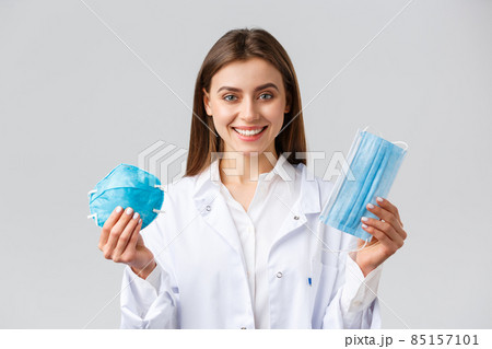 Covid-19, preventing virus, healthcare workers concept. Smiling attractive female doctor in white scrubs showing two variants of personal protective equipment, show respirator and medical mask 85157101