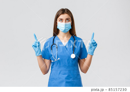 Covid-19, preventing virus, health, healthcare workers and quarantine concept. Curious female doctor or nurse in blue scrubs, medical mask reading banner, pointing fingers up Covid-19, preventing virus, health, healthcare workers and quarantine concept. Curious female doctor or nurse in blue scrubs, medical mask reading banner, pointing fingers up 85157230