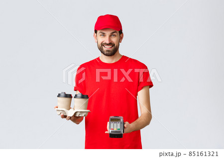 Food delivery, quarantine, stay home and order online concept. Friendly bearded courier in red uniform handing coffee delivery and POS terminal for client to pay contactless, grey background 85161321