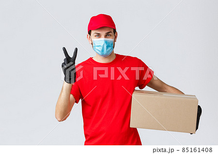 Packages and parcels delivery, covid-19 quarantine delivery, transfer orders. Friendly courier in red uniform, face mask and gloves, deliver order to client, holding box, show peace sign Packages and parcels delivery, covid-19 quarantine delivery, transfer orders. Friendly courier in red uniform, face mask and gloves, deliver order to client, holding box, show peace sign 85161408