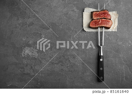 Carving fork with slices of delicious beef steak on grey table, top view. Space for text Carving fork with slices of delicious beef steak on grey table, top view. Space for text 85161640