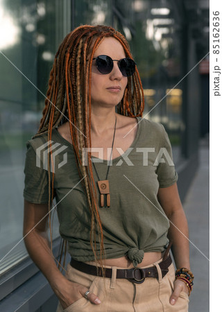 Young beautiful girl with red dreadlocks and glasses stands leaning against the building. Street portrait of young woman 85162636