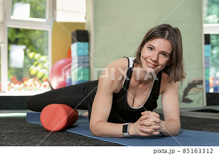 Young athletic smiling girl kneads her hip muscles with equipment for self-masage. rollers. Myofascial release with foam roll. Young athletic smiling girl kneads her hip muscles with equipment for self-masage. rollers. Myofascial release with foam roll. 85163312