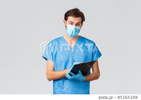 Covid-19, quarantine, hospitals and healthcare workers concept. Cheerful doctor in medical mask, scrubs and gloves, looking at patient, listening to problem and symptoms, writing down at clipboard Covid-19, quarantine, hospitals and healthcare workers concept. Cheerful doctor in medical mask, scrubs and gloves, looking at patient, listening to problem and symptoms, writing down at clipboard 85165109