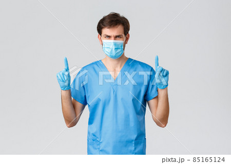 Covid-19, healthcare workers and hospital concept. Serious-looking male doctor in blue scrubs, medical mask and gloves, pointing fingers up, showing you important sign or news, top advertisement Covid-19, healthcare workers and hospital concept. Serious-looking male doctor in blue scrubs, medical mask and gloves, pointing fingers up, showing you important sign or news, top advertisement 85165124