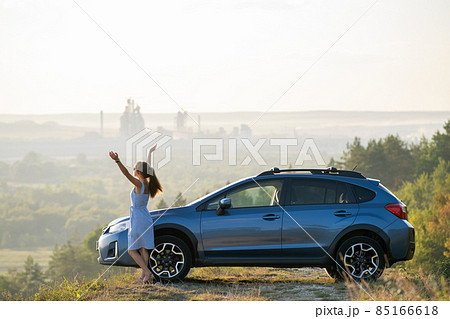 Young female driver resting near her car raising up her hands on warm summer evening. Travel destinations concept. 85166618