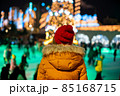 Young woman in red hat and yellow jacket looking at people skating on ice rink near blurred Christmas tree 85168715