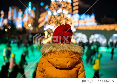 Young woman in red hat and yellow jacket looking at people skating on ice rink near blurred Christmas tree 85168715
