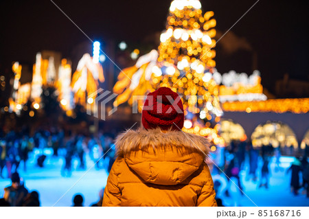 Young woman in red hat and yellow jacket looking at people skating on ice rink near blurred Christmas tree 85168716
