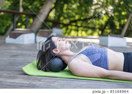 Athletic girl lies on yoga mat and does neck massage with myofascial balls. Kneads the muscles of neck with MFR roller outdoors 85168964