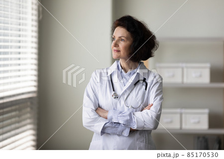 Smiling pensive older female doctor standing by window in office 85170530