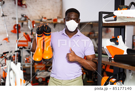 Man wearing protective mask chooses rubber boots at garden supply store 85177140
