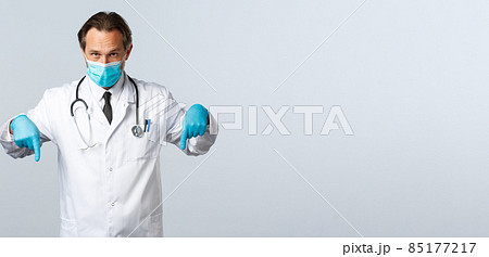 Covid-19, preventing virus, healthcare workers and vaccination concept. Serious doctor in medical mask and gloves, pointing fingers down, showing promo banner, white background Covid-19, preventing virus, healthcare workers and vaccination concept. Serious doctor in medical mask and gloves, pointing fingers down, showing promo banner, white background 85177217