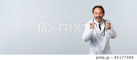 Covid-19, coronavirus outbreak, healthcare workers and pandemic concept. Carefree and happy male doctor laughing over funny joke, pointing at camera and chuckle, white background 85177499