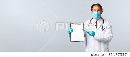 Covid-19, preventing virus, healthcare workers and vaccination concept. Shocked male doctor in medical mask and gloves pointing at clipboard, stare astonished, showing important information 85177537