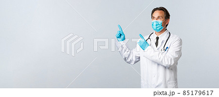 Covid-19, preventing virus, healthcare workers and vaccination concept. Middle-aged male doctor in white coat, medical mask and gloves looking upper left corner at pointing banner 85179617