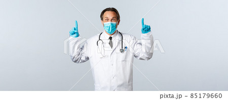 Covid-19, preventing virus, healthcare workers and vaccination concept. Impressed surprised male doctor in medical mask and gloves, pointing fingers up to show great news Covid-19, preventing virus, healthcare workers and vaccination concept. Impressed surprised male doctor in medical mask and gloves, pointing fingers up to show great news 85179660