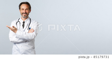 Covid-19, coronavirus outbreak, healthcare workers and pandemic concept. Handsome friendly physician showing way. Doctor in white coat pointing hand left at promo, inviting patients 85179731