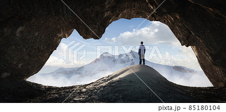 Adult Hiker Male standing inside a rocky mountain cave overlooking the nature scene 85180104