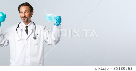 Covid-19, preventing virus, healthcare workers and vaccination concept. Pleased doctor in gloves and white coat showing medical respirator and mask, explain importance wearing PPE during coronavirus 85180563