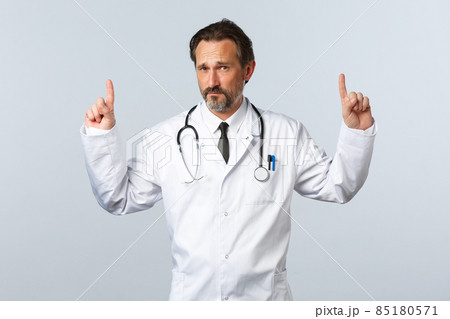Covid-19, coronavirus outbreak, healthcare workers and pandemic concept. Skeptical adult male doctor in white coat pointing fingers up and looking concerned at camera, have doubts 85180571