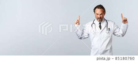 Covid-19, coronavirus outbreak, healthcare workers and pandemic concept. Angry outraged male doctor in white coat, pointing fingers up grimacing furious, looking mad at camera, white background 85180760