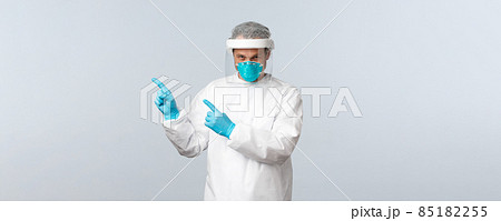 Covid-19, preventing virus, healthcare workers and vaccination concept. Serious doctor in personal protective equipment pointing fingers upper left corner at information banner Covid-19, preventing virus, healthcare workers and vaccination concept. Serious doctor in personal protective equipment pointing fingers upper left corner at information banner 85182255