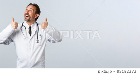 Covid-19, coronavirus outbreak, healthcare workers and pandemic concept. Enthusiastic handsome doctor in white coat looking at top left advertisement and thumbs-up, recommend product 85182272