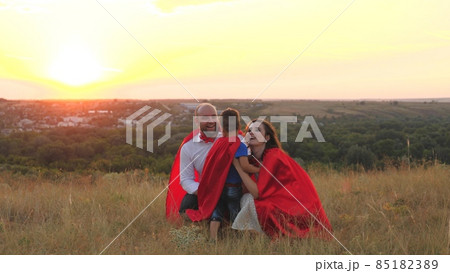 mother and father kiss and hug little child sunset, playing superheroes dressed red cloaks, happy family, brave savior costume, fantastic childhood dream heroes, fearless kid boy winner fun play game 85182389