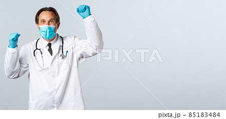 Covid-19, preventing virus, healthcare workers and vaccination concept. Relieved supportive doctor in medical mask and gloves, raising hands up as chanting, celebrating achievement of goal 85183484