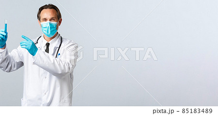 Covid-19, preventing virus, healthcare workers and vaccination concept. Satisfied smiling doctor in medical mask and gloves pointing at test-tube with vaccine sample, approve medication Covid-19, preventing virus, healthcare workers and vaccination concept. Satisfied smiling doctor in medical mask and gloves pointing at test-tube with vaccine sample, approve medication 85183489