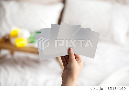 Hand of unknown person holding some empty pieces of paper against blurred bed with white linen and breakfast serving tray on it. Close up, copy space 85184169