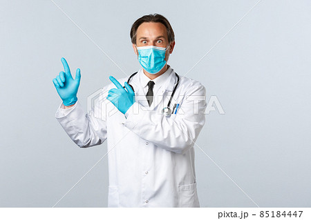 Covid-19, preventing virus, healthcare workers and vaccination concept. Surprised startled doctor in white coat, medical mask and gloves pointing upper left corner astonished, showing advertisement 85184447
